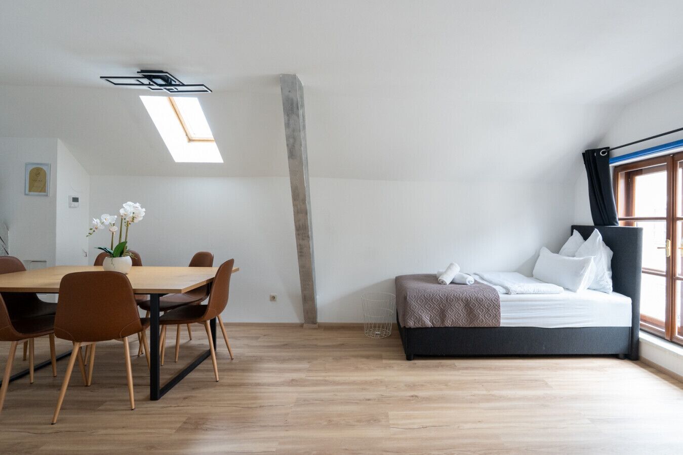 A modern room with a wooden dining table, chairs, a bed with a headboard, and a skylight.