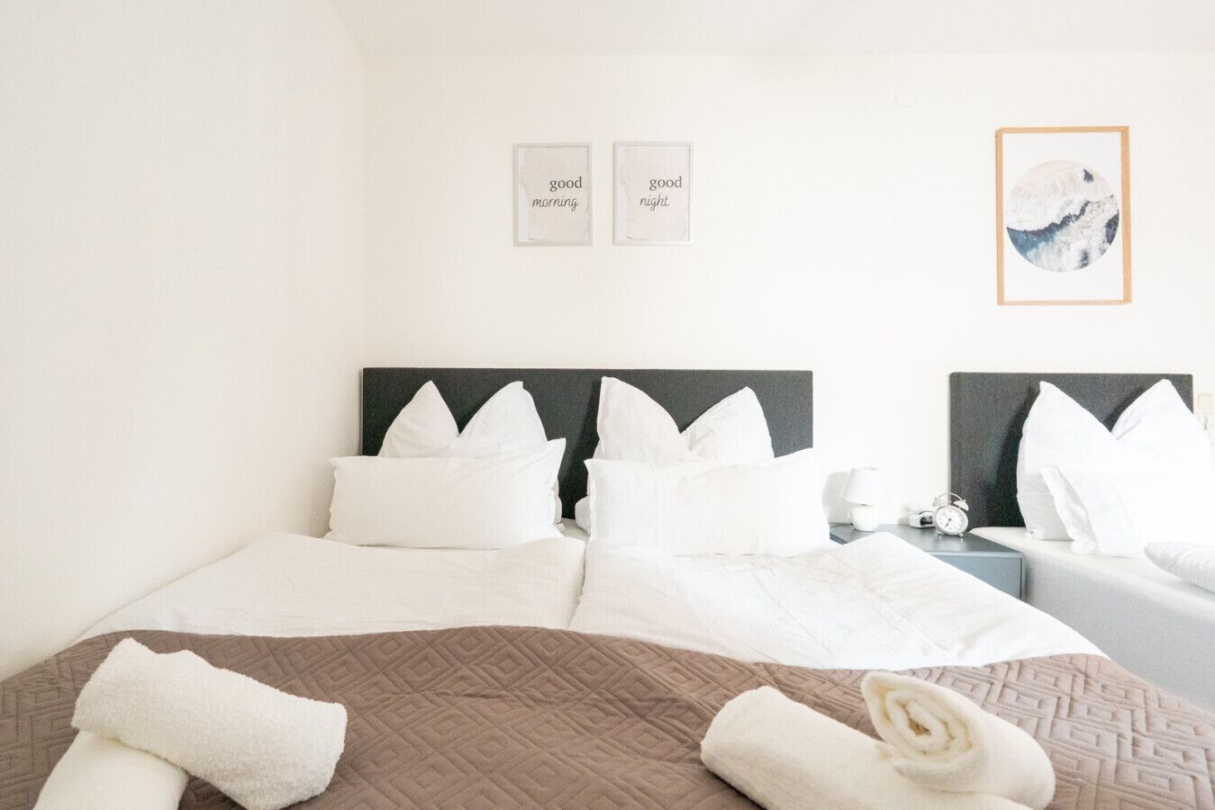 Een hotelkamer met twee bedden, wit beddengoed, een bruine sprei, twee nachtkastjes met lampen, en ingelijste foto’s aan de muur.