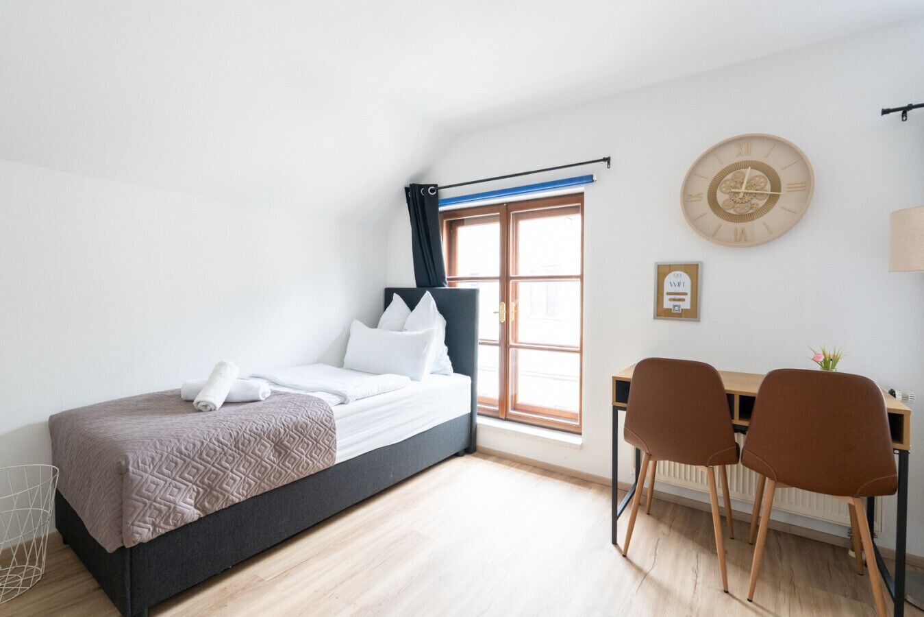 Een slaapkamer met een bed, een bureau, twee stoelen, een raam met gordijnen, een wandklok en een radiator.