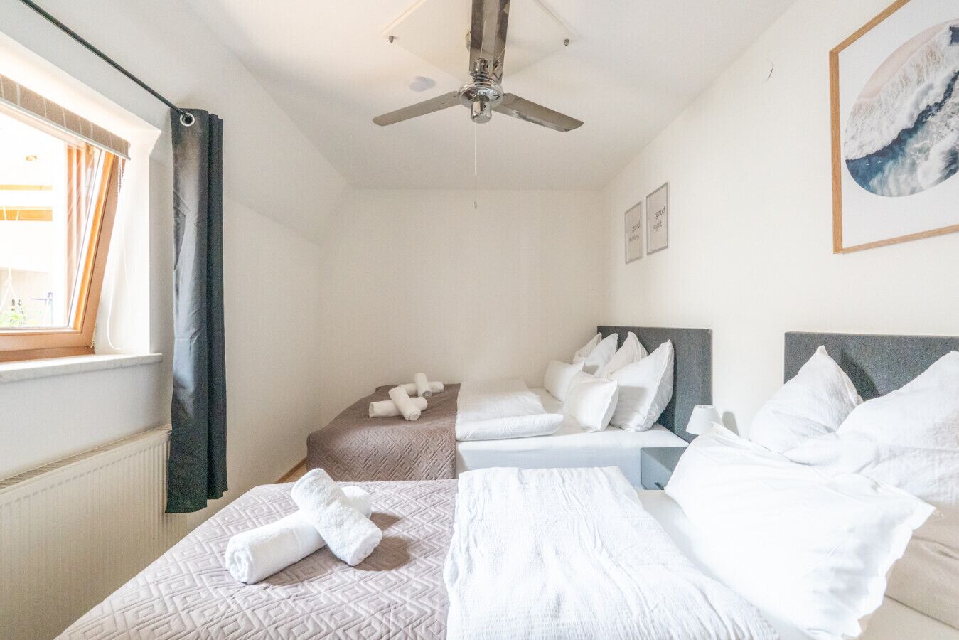 Ett hotellrum med två sängar, en takfläkt, ett fönster med gardiner och en inramad tavla på väggen.