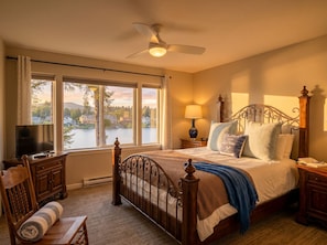 A bedroom with a bed, a TV, a desk, a chair, a window with a view of a lake and houses, and a ceiling fan.