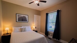 A hotel room with a bed, a ceiling fan, a window with curtains, a nightstand with a lamp, and a framed picture on the wall. - College St. Cottage just 10 minutes from WEC. (Wilmington)