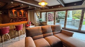 A cozy bar area with a sofa and stools, large windows with a view of the outdoors, and a stone wall.