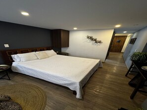 A hotel room with a large bed, a bedside table, a lamp, a plant, and a wooden floor. - Lourdes Executive Suites 201 (Bogotá)