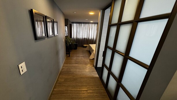 A hallway with wooden flooring, a bed with a beige cover, and a large window with a grid pattern. - Lourdes Executive Suites 201 (Bogotá)