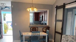 Un comedor con una mesa y sillas, una cocina al fondo, un sillón estampado y una puerta corredera.