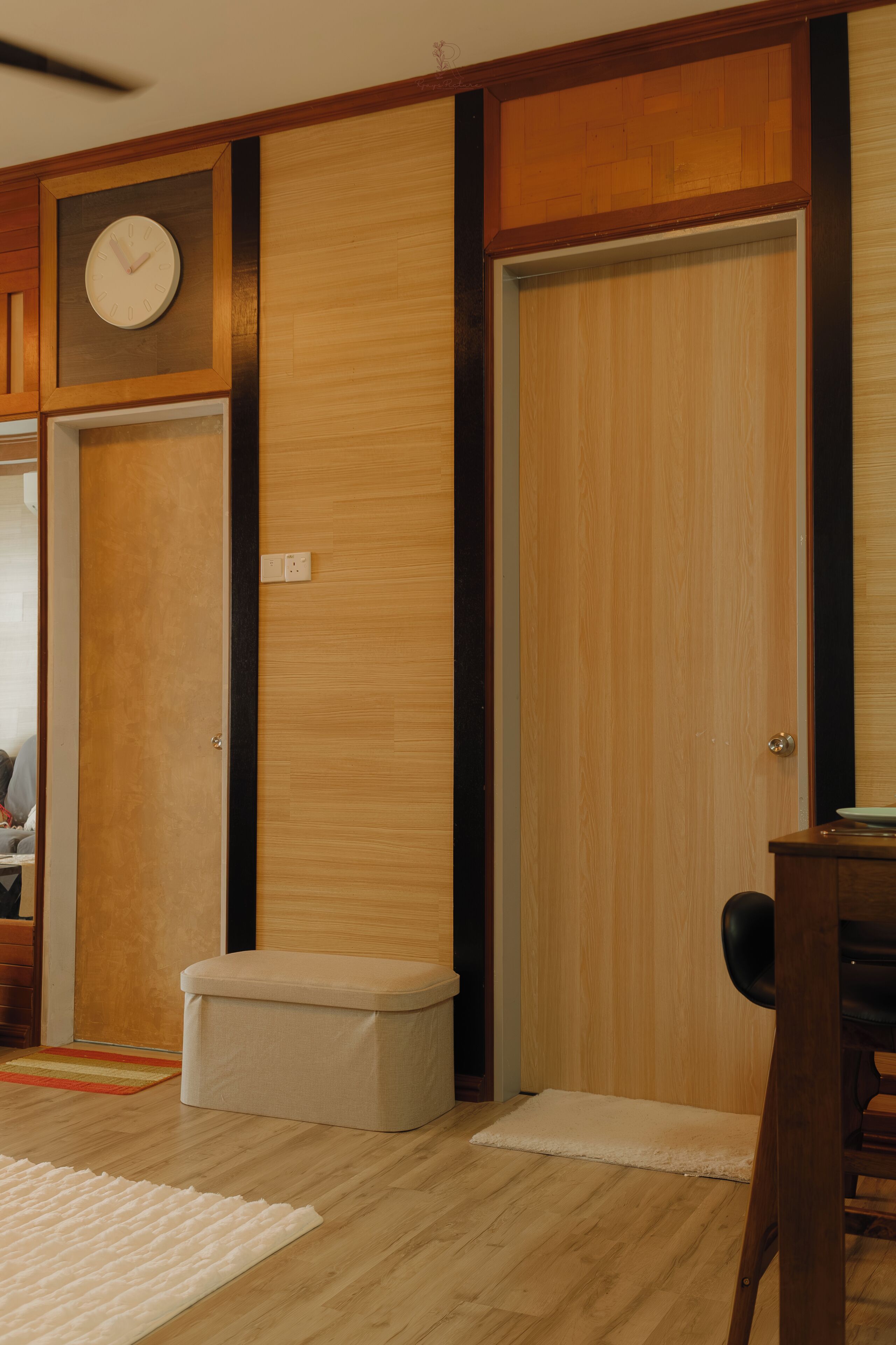 A room with a wooden floor, a white rug, a wooden bench, a small stool, and a door with a clock on the wall.