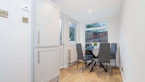 Cocina compacta con armarios blancos, una mesa de comedor con cuatro sillas y vista a un jardín a través de una ventana.