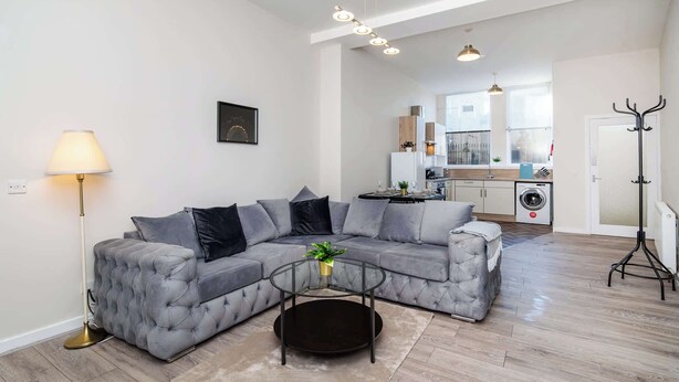A modern living room with a grey sectional sofa, a glass coffee table, and a floor lamp. A kitchen with a washing machine is visible in the background. - Parking Aberdeenshire Retreat Central Peterhead (Aberdeenshire)