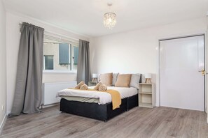 A bedroom with a bed, a radiator, a window with curtains, a door, and a chandelier.