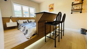 A compact room with a bed, a desk, two chairs, a small shelf with decorative items, and a window.