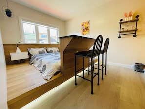 Habitación pequeña con cama, escritorio, dos sillas, un estante con adornos y una ventana.