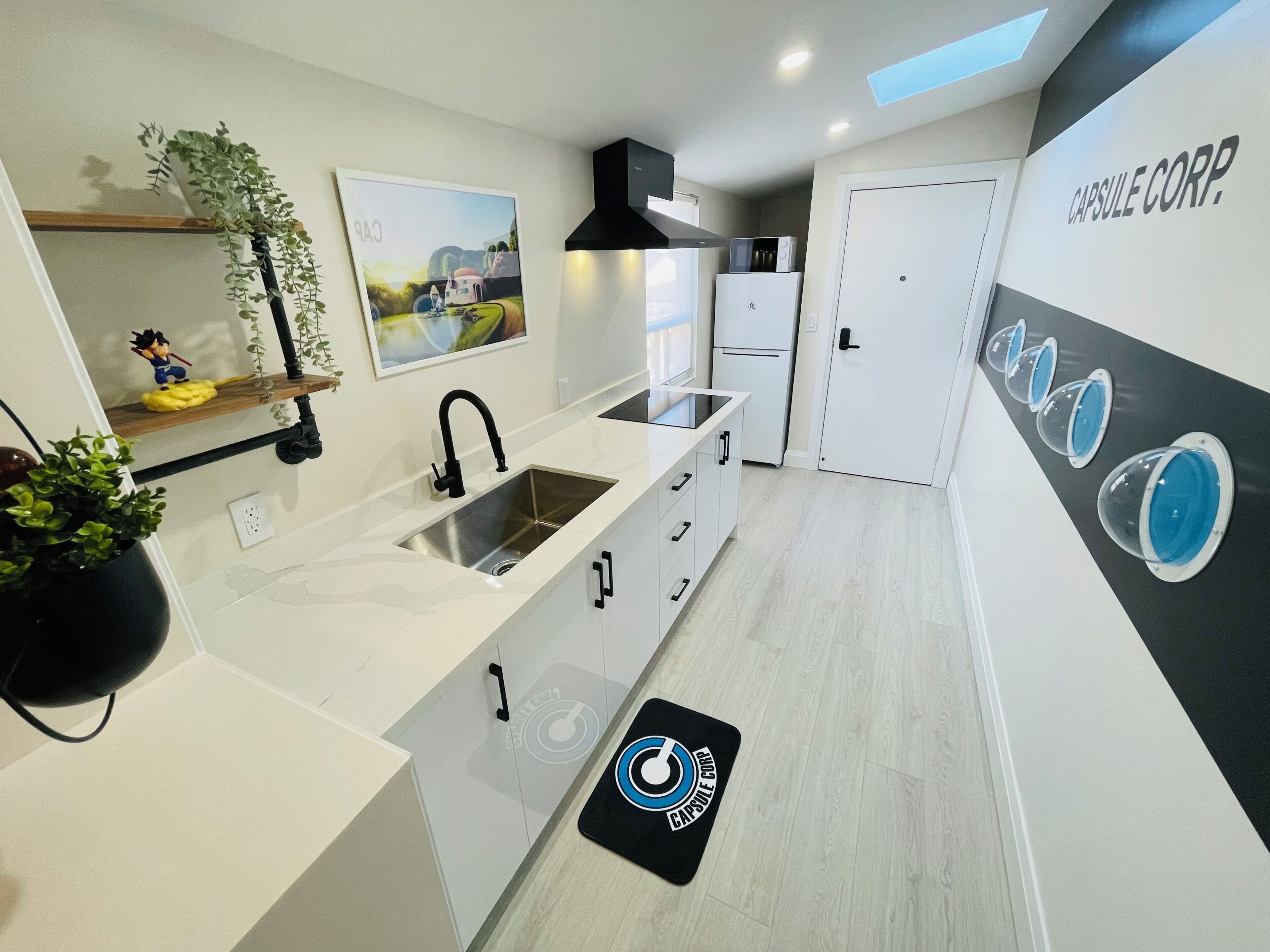 Una cocina moderna con un lavamanos, armarios, un microondas y un refrigerador. Hay una planta en la encimera y una puerta con un logotipo.