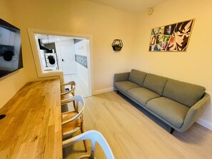 Una sala de estar con sofá, una mesa de comedor con sillas, un televisor montado en la pared y una puerta que da a la cocina.