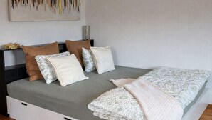 A bedroom with a bed, pillows, a blanket, a wall-mounted light, and a painting.