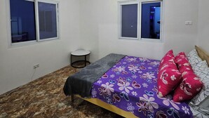 Een slaapkamer met een bed, twee ramen, een klein nachtkastje en een plafondlamp.