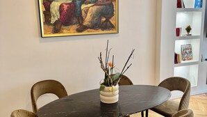 A dining area with a round table, six chairs, a painting on the wall, and a bookshelf with books and decorative items. - Luxury Apartment Paris Trocadero- 2 bedrooms 80m2 (Paris)