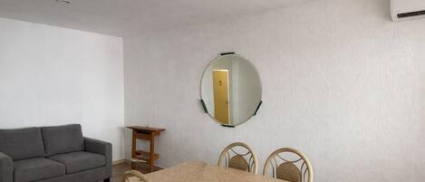 Une pièce avec un canapé gris, une table en bois avec quatre chaises, un miroir rond accroché au mur et un appareil de climatisation.