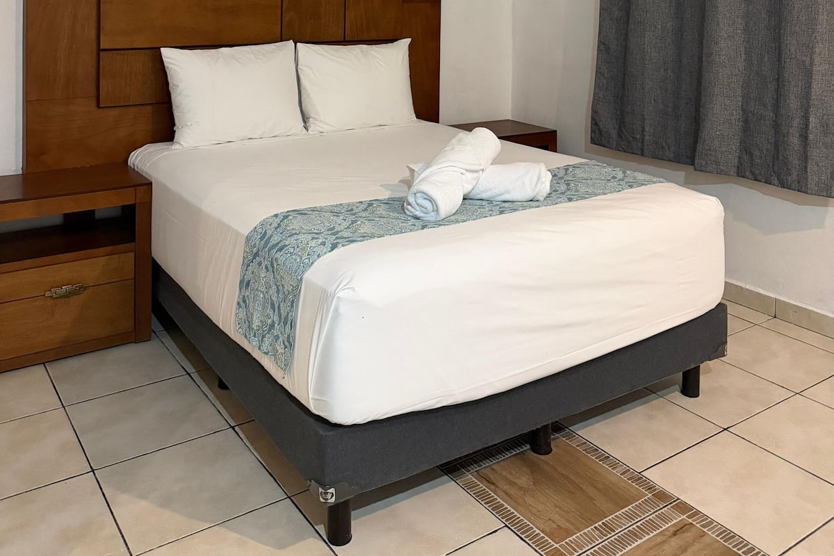 Una habitación de hotel con cabecera de madera, una cama con ropa blanca, una mesita de noche y una ventana con cortinas grises.