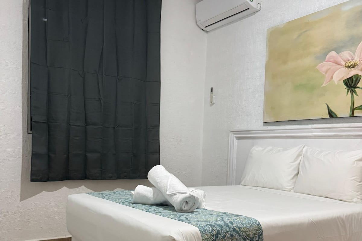 Habitación de hotel con cama, ventana con cortinas oscuras, unidad de aire acondicionado y un cuadro de una flor en la pared.
