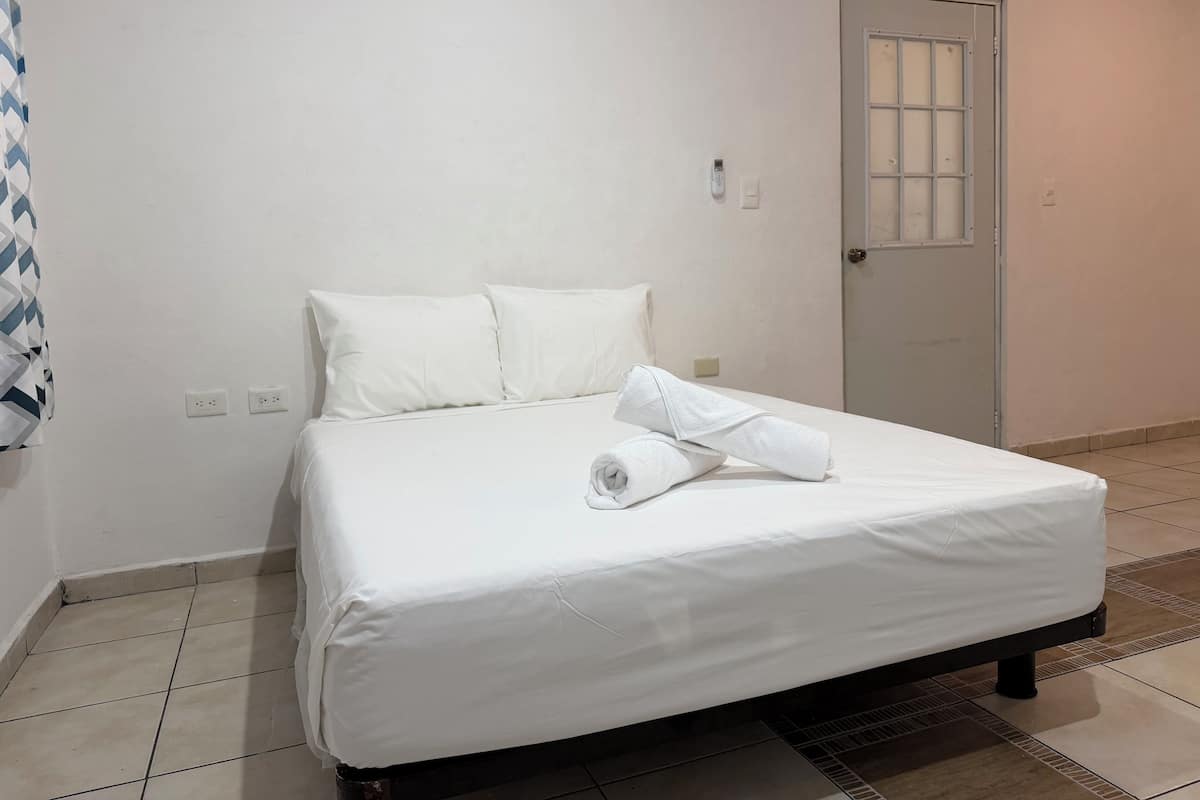 Una cama bien tendida con sábanas blancas, una puerta con ventanal y una pared blanca sin adornos con dos tomas de corriente.