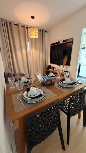 A dining area with a wooden table set for four, a television mounted on the wall, and a sofa in the background.