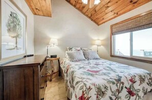 Ein Schlafzimmer mit Holzboden, einem Bett mit floraler Bettwäsche, Nachttischlampen, einem Nachttisch und einem gerahmten Bild an der Wand.