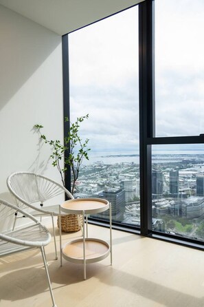 A modern room with a large window, a white chair, a small round table, and a potted plant. - Stunning Harbour View | 2BR 2BA Suite (Melbourne)