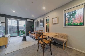 A modern living room with a round table, two chairs, a sofa, and a bench. There is a large window with a view of trees and a door leading to a balcony. - Cozy & Stylish Central City Retreat (Christchurch)