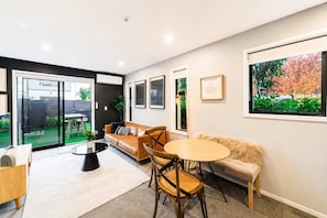 A modern living room with a sofa, chairs, a coffee table, and a large window with a view of a garden. - Central City Stay | 1BR | Work Space + Courtyard (Christchurch)