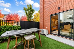 Eine Terrasse mit Holztisch und -stühlen, einem schwarzen Zaun und einem Gebäude mit Glastür, die zu einem Innenbereich führt.