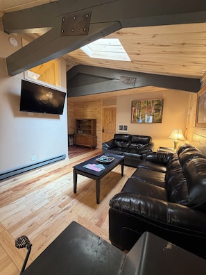Ein Wohnzimmer mit schwarzem Ledersofa, Couchtisch, Flachbildfernseher, Holzboden und einem Oberlicht.