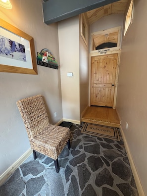 A room with a stone-patterned floor, a wicker chair, a wooden door labeled 'Box Canyon Retreat,' and a welcome mat. - The Box Canyon Retreat on Main St. with AC! (Ouray)