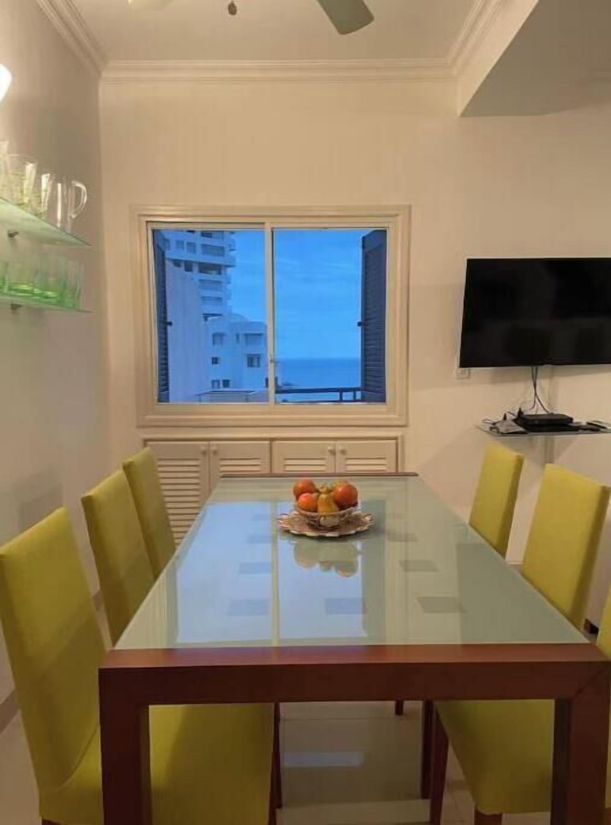 Un coin repas avec une table en verre, des chaises jaunes, un saladier de fruits, un téléviseur et une vue sur les bâtiments par la fenêtre.