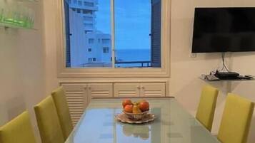 Un coin repas avec une table en verre, des chaises jaunes, un saladier de fruits, un téléviseur et une vue sur les bâtiments par la fenêtre.