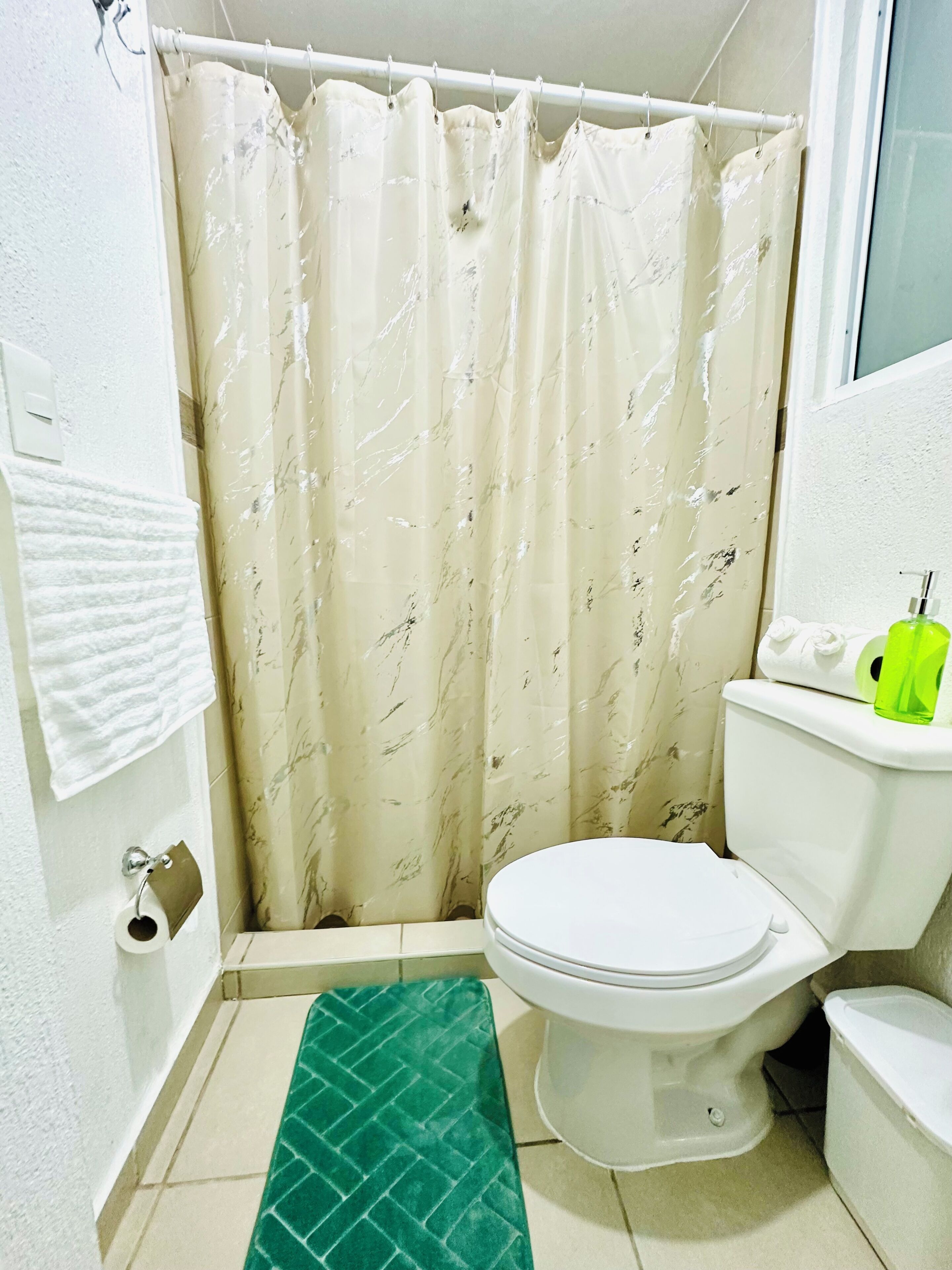 A bathroom with a toilet, a green mat, a shower curtain, and a green bottle on the toilet tank.