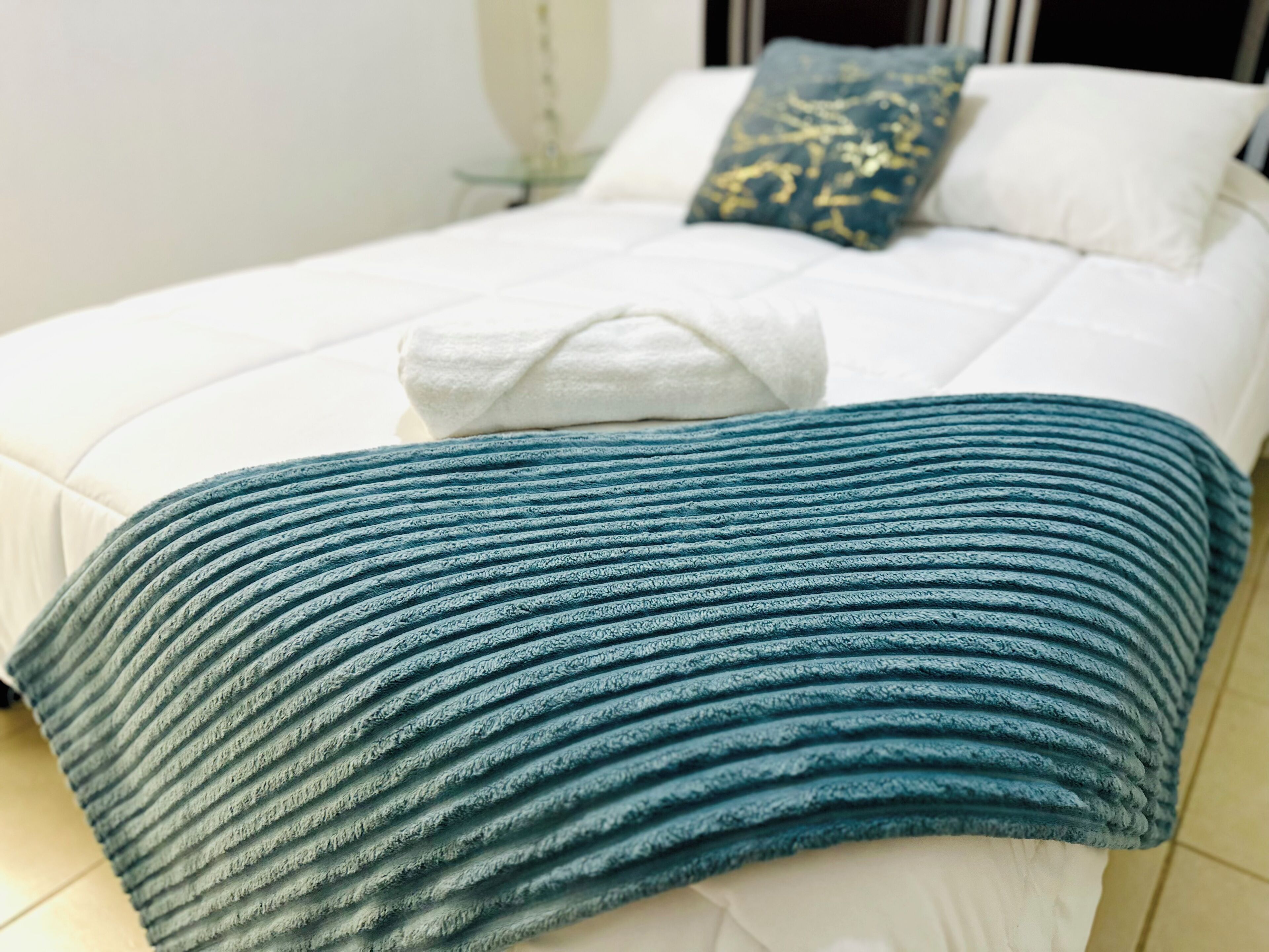 A bed with a white mattress, a teal textured blanket, and a blue pillow with gold patterns.