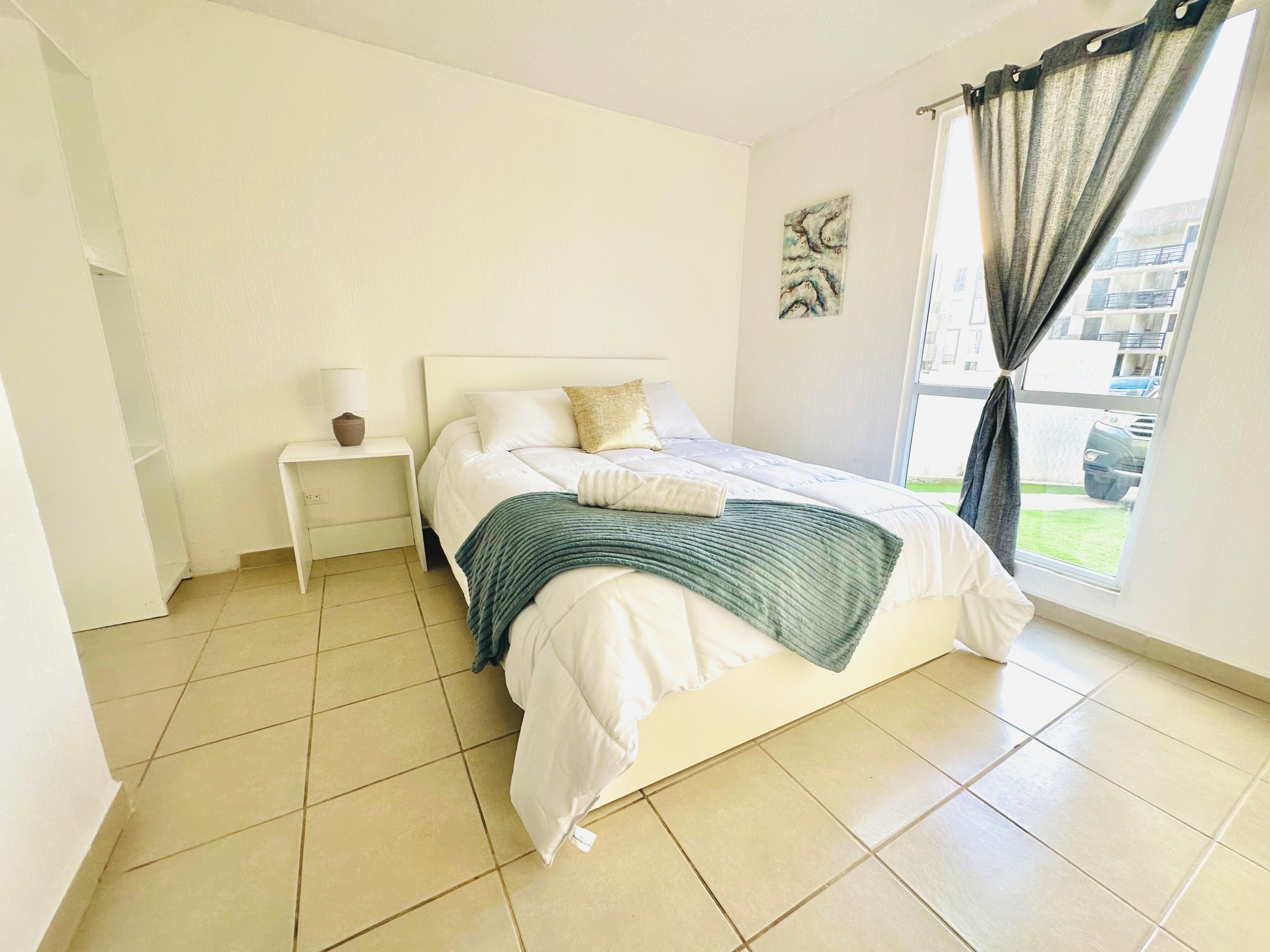 Een slaapkamer met een bed, een nachtkastje, een lamp, een raam met gordijnen en een schilderij aan de muur.