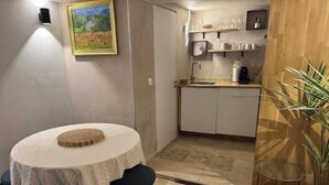 Una cocina compacta con armarios blancos, un lavamanos y una encimera. Hay una mesa redonda con mantel blanco y dos sillas. Un cuadro adorna la pared, y una planta en maceta está a la derecha.