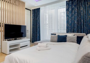 A modern hotel room with a flat-screen TV, a sofa bed, a TV stand, and a large window with curtains. - Tauron Arena Chic Apartment (Kraków)