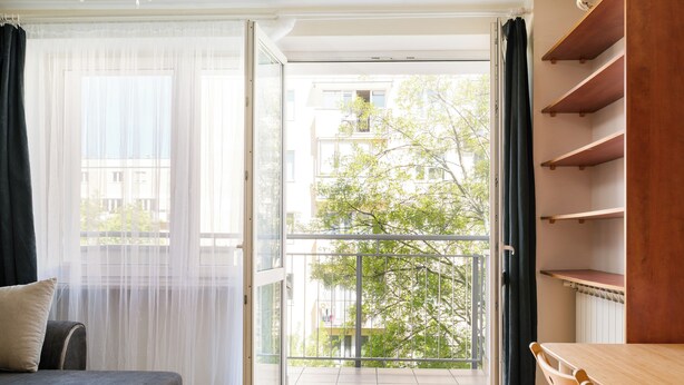 Ein Zimmer mit einer Couch, einem Esstisch mit Stühlen, einem Balkon mit Blick auf Gebäude und Bäume sowie einem Fenster mit durchsichtigen Vorhängen.