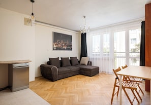 Un salon moderne avec un parquet en chevrons, un canapé d’angle, une table à manger avec des chaises et un lave-vaisselle.