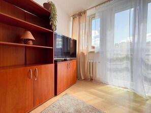 Ein Zimmer mit Holzausstattung, einem Fernseher, einem Topfpflanzen, einem Heizkörper und einem großen Fenster mit durchsichtigen Vorhängen.