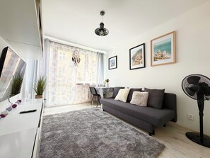 A modern living room with a grey sofa, a large window, a dining table, and a ceiling fan. - Reja Beige Apartment (Wrocław)