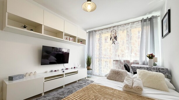 Une chambre moderne avec une télévision, des meubles blancs, une grande fenêtre avec des rideaux et un lit recouvert d’une couverture beige.