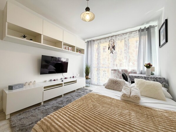 A modern bedroom with a TV, white furniture, a large window with curtains, and a bed with a beige blanket. - Reja Beige Apartment (Wrocław)