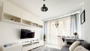 Un salon moderne doté d’un téléviseur à écran plat, d’un meuble de rangement blanc, d’un canapé gris et d’une grande fenêtre agrémentée de rideaux légers.