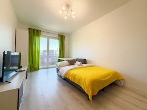A bedroom with a bed, a desk with a computer, a window with curtains, and a ceiling light fixture.