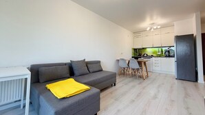 A modern living room with a grey sofa, a small white table, and a yellow throw blanket. A kitchen with white cabinets and a black refrigerator is visible in the background.