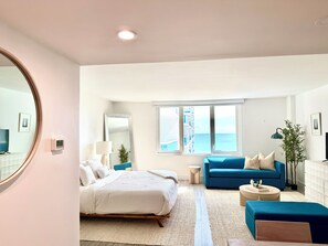 Ein modernes Schlafzimmer mit Bett, einem blauen Sofa, einem kleinen runden Tisch und einem großen Fenster mit Blick auf das Meer.
